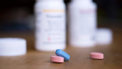 Vor zwei geöffneten Tablettendosen liegen zwei rosane und eine blaue Tablette.